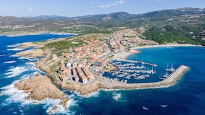 An aerial view of a coastal town with a marina, rocky coastline, and surrounding hills. - Cozy Accommodation With Shared Pool (Aglientu)
