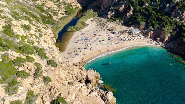An aerial view of a coastal beach with rocky cliffs, clear turquoise waters, and a sandy beach area with umbrellas and people. - Cozy Accommodation With Shared Pool (Aglientu)