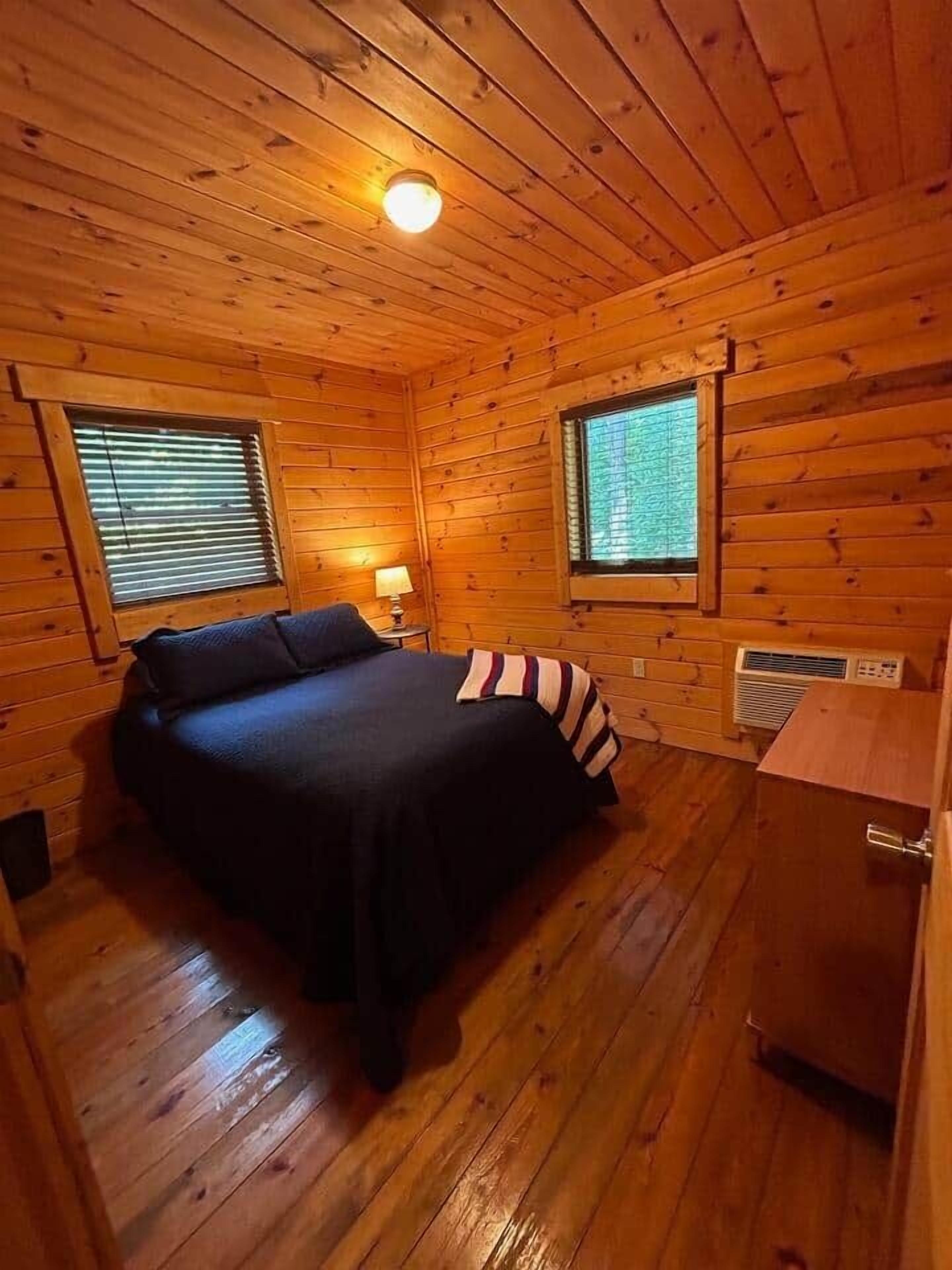 Una habitación pequeña y acogedora con paredes de madera y una cama. Hay dos ventanas con persianas, un calentador y una mesita con una lámpara.