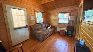 A cozy cabin interior with wooden walls, a brown leather sofa, a door with blinds, and a small heater. - Justin Fitch Cabin - Active Heroes (Bullitt County)