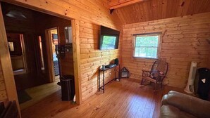 A log cabin interior with a wooden floor, a television mounted on the wall, a rocking chair, and a small table with a trash can. - Justin Fitch Cabin - Active Heroes (Bullitt County)