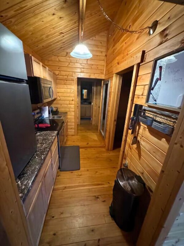 A narrow wooden hallway with a kitchen area on one side, including a refrigerator, microwave, and countertop.