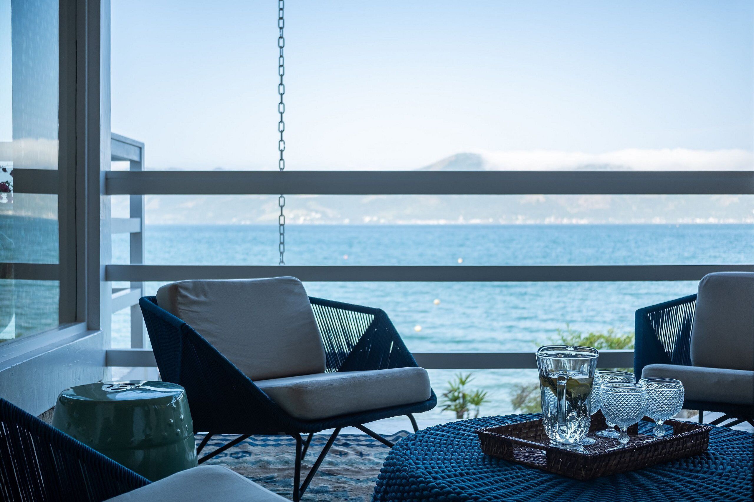 A balcony with a view of the sea, featuring two chairs, a table with glasses, and a decorative stool.