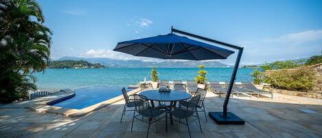 A poolside area with a table and chairs under a large umbrella, overlooking a body of water and mountains.