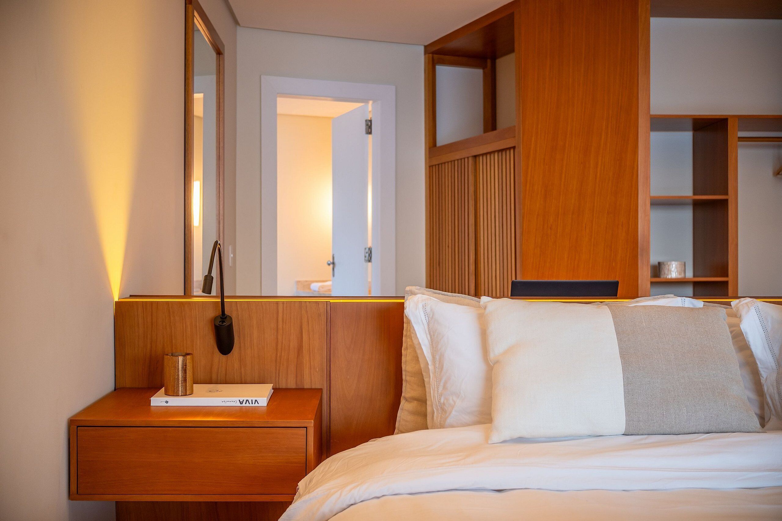A hotel room with a wooden nightstand, a bedside table, a bed with white bedding, and a wooden wardrobe with shelves.