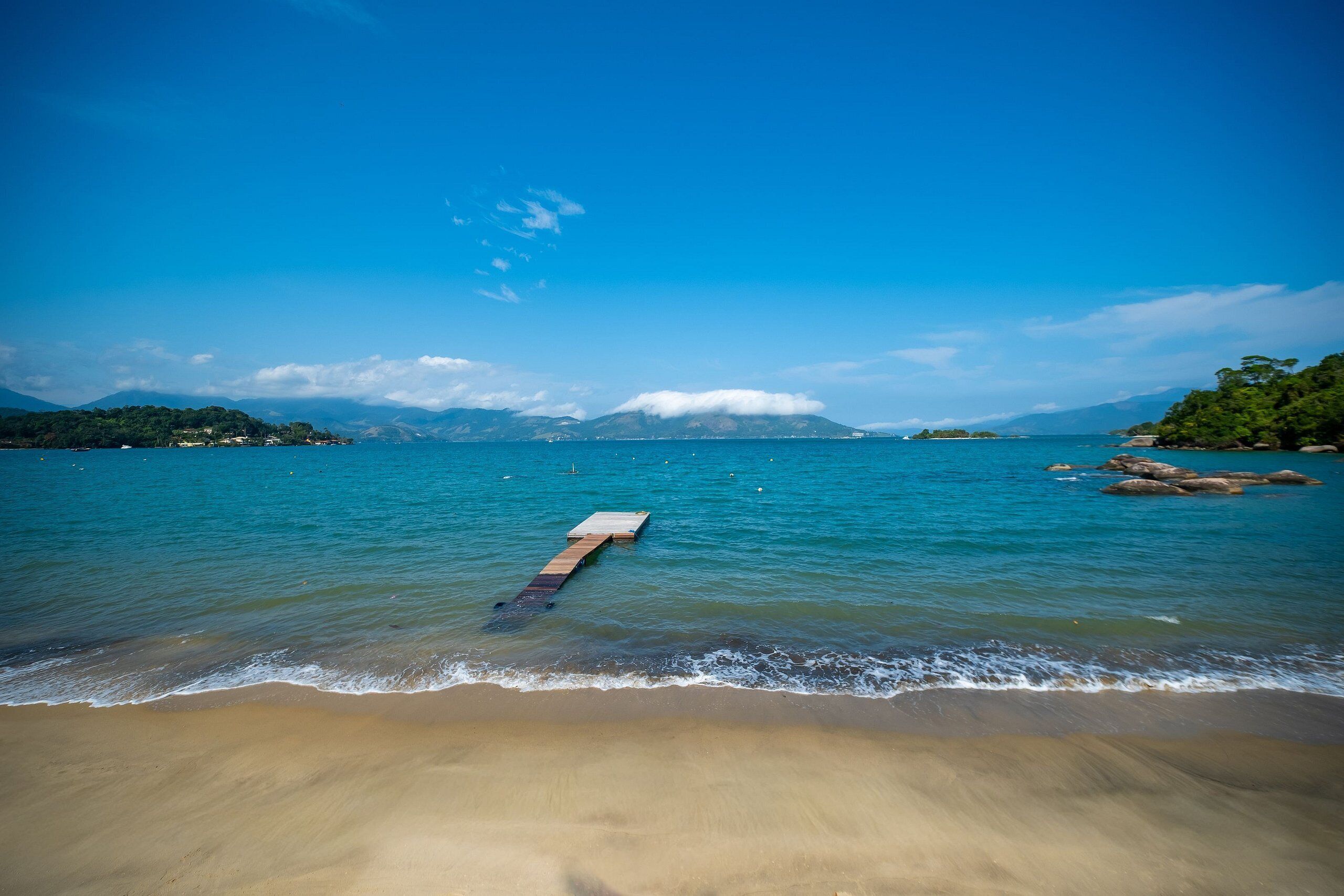 Una playa con un muelle de madera que se extiende sobre aguas tranquilas, rodeada de montañas y cielos despejados.