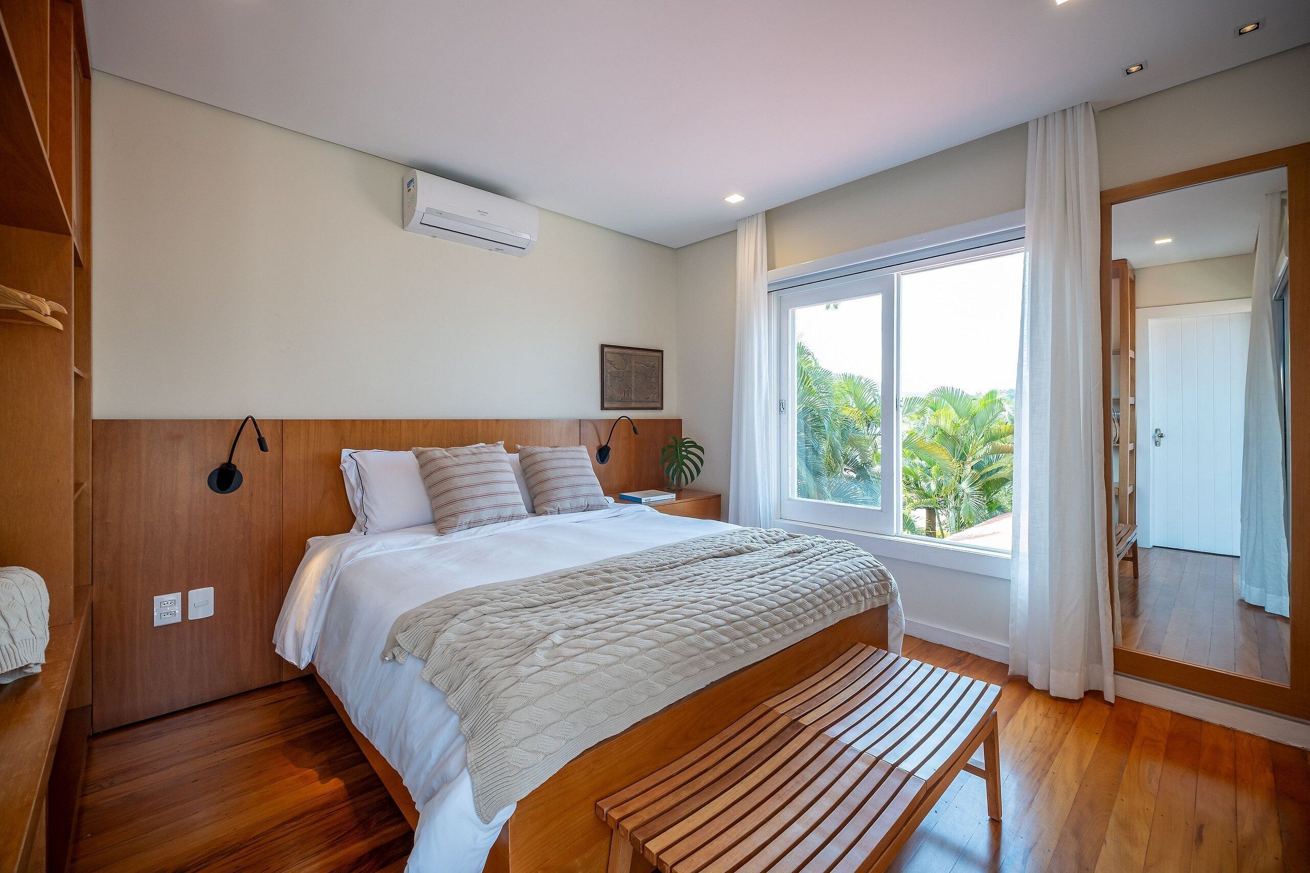 A modern bedroom with a wooden bed, a large window with a view of palm trees, and a wooden bench.