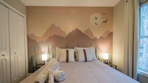 A bedroom with a mountain mural, two bedside lamps, a neatly made bed, and a window with a view. - Spacious 4BR Retreat w/Free Tix | Fire Pit + Grill (Asheville)