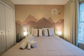 Ein Schlafzimmer mit Wandgemälde von Bergen, zwei Nachttischlampen, einem ordentlich bezogenen Bett und einem Fenster mit Aussicht.