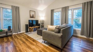A living room with a gray sofa, a TV, and a wooden floor. - Spacious 4BR Retreat w/Free Tix | Fire Pit + Grill (Asheville)