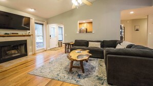 A spacious living room with a fireplace, a large flat-screen TV, a ceiling fan, and a view of the outdoors. - Spacious 4BR Retreat w/Free Tix | Fire Pit + Grill (Asheville)