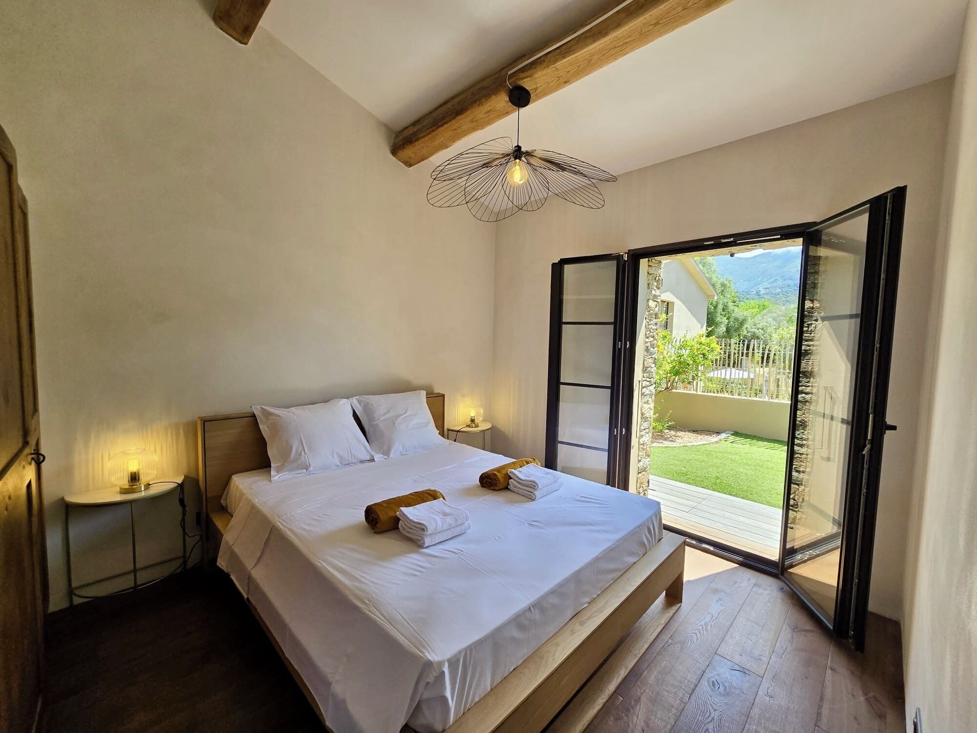 Een slaapkamer met een bed, bedlampjes en uitzicht op een tuin door een open deur.