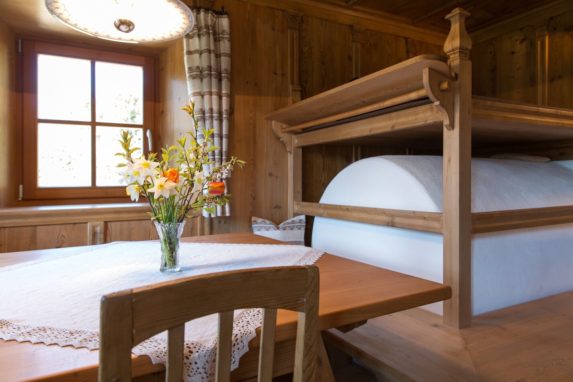 A wooden bunk bed with a white mattress, a wooden table with a lace tablecloth, and a vase with flowers.