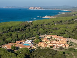 An aerial view of a coastal residential area with houses, a swimming pool, and a clear view of the sea and distant mountains. - Cozy Flat in a Residence With Pool (Aglientu)