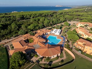An aerial view of a resort with a swimming pool surrounded by villas, a clear view of the sea, and a lush green landscape. - Cozy Flat in a Residence With Pool (Aglientu)