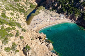 Vista aérea de uma praia costeira com falésias rochosas, águas turquesas cristalinas e uma área de areia com guarda-sóis e pessoas.