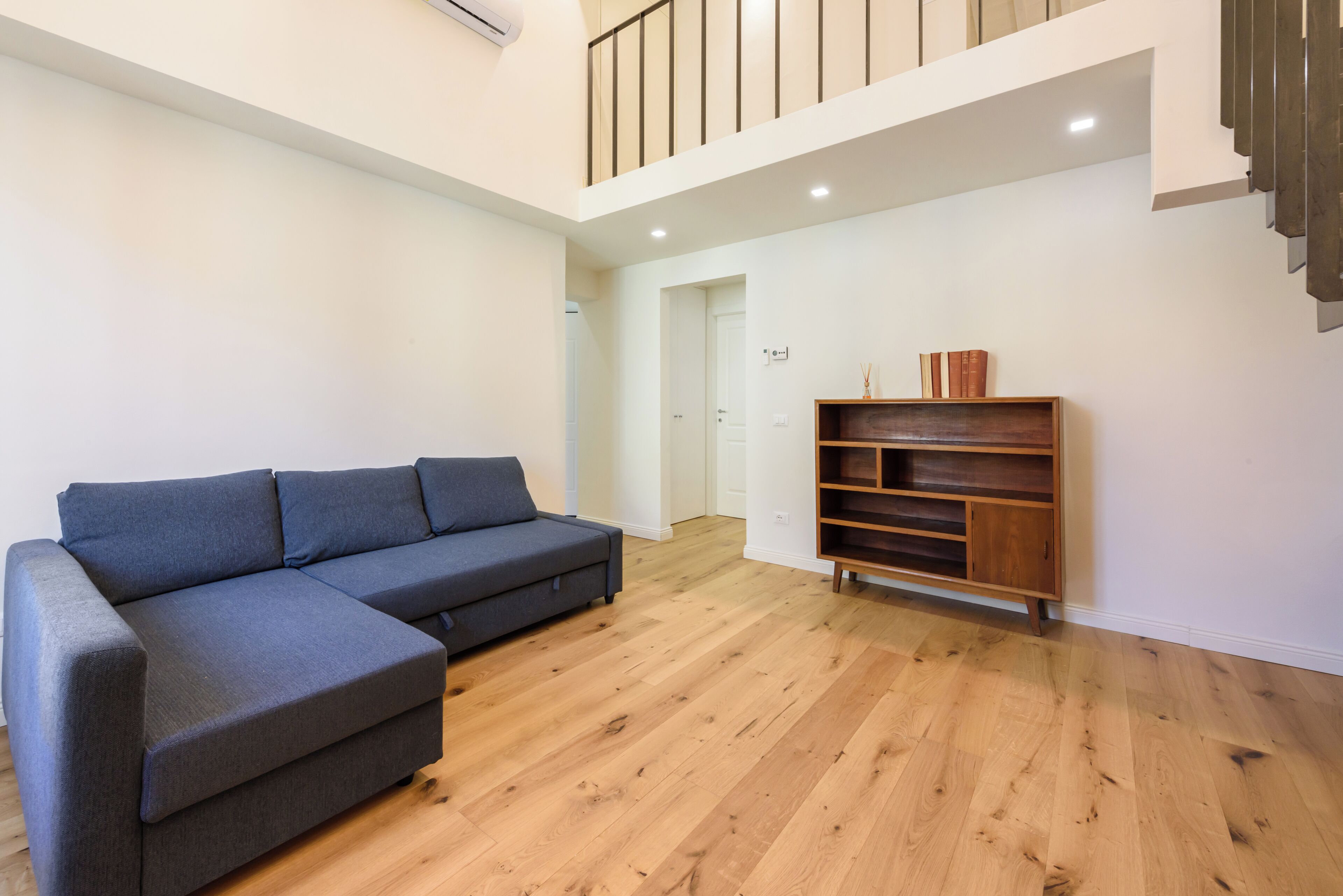A modern living room with a sectional sofa, wooden flooring, a built-in bookshelf, and a staircase leading to an upper level.