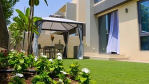 A modern outdoor patio with a covered seating area, a well-maintained lawn, and a variety of plants. - 4BR + Maids Villa in Maple 2, Dubai Hills (Dubai)