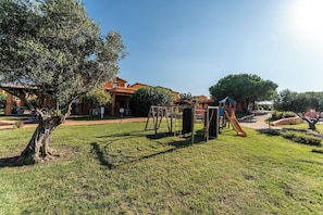 A playground with swings and a slide, surrounded by grass and trees, with buildings in the background. - Cozy Studio With Shared Pool (Aglientu)