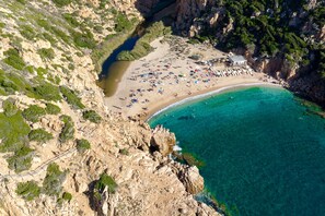 An aerial view of a coastal beach with rocky cliffs, clear turquoise waters, and a sandy beach area with umbrellas and people. - Cozy Studio With Shared Pool (Aglientu)