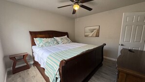 A bedroom with a wooden bed, a small bedside table, a ceiling fan, a door, and a framed picture on the wall.