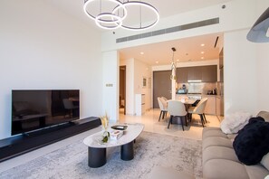 Una sala de estar moderna con televisor de pantalla plana, una mesa de centro con objetos decorativos, un sofá con una manta y un comedor con sillas, con una cocina al fondo.