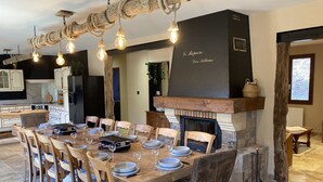 A dining area with a long wooden table set for six, surrounded by chairs. A fireplace with a stone mantle is visible in the background. - Maison Auvergnate Le repaire des Milans (Laveissière)