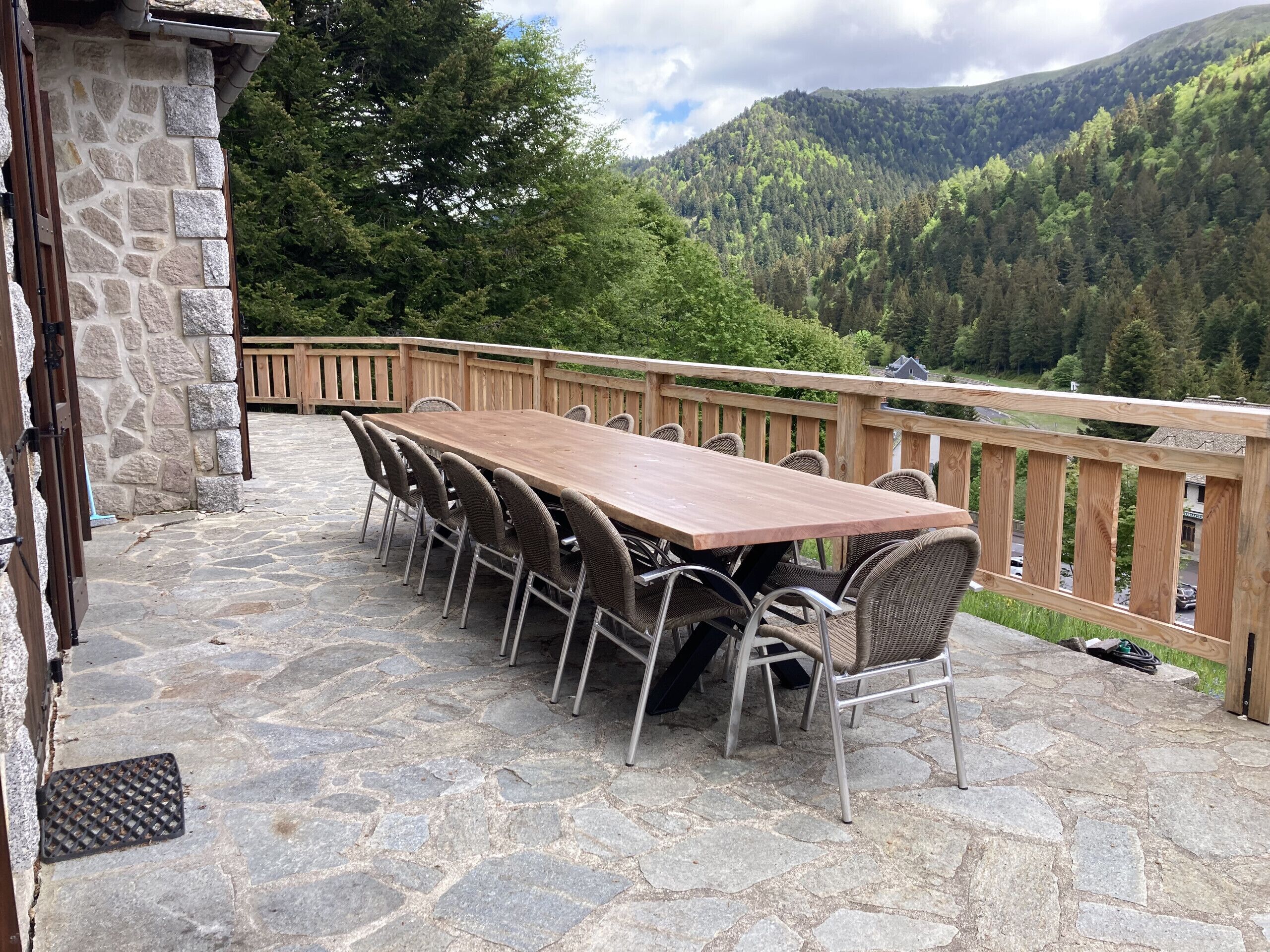 A stone patio with a wooden deck, a long table with chairs, and a view of a forested mountain.