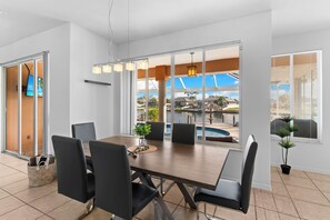 Un espace de repas moderne, doté d’une grande table et de chaises, de larges fenêtres offrant une vue sur une piscine et un espace extérieur, ainsi que d’un plafond agrémenté de luminaires suspendus.