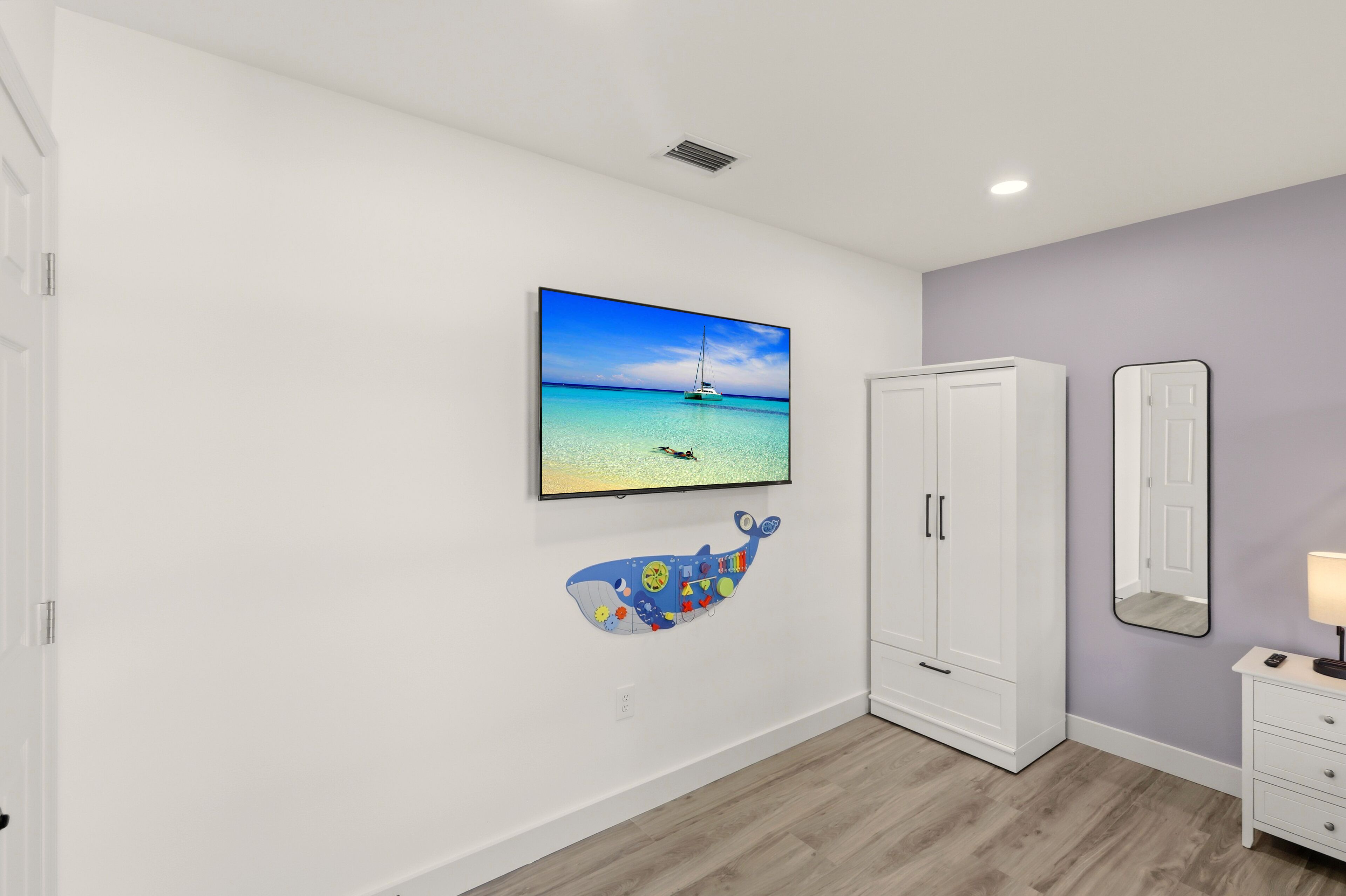 Ein Zimmer mit einem weißen Kleiderschrank, einem an der Wand befestigten Fernseher, der ein Segelboot auf dem Meer zeigt, und einer bunten Wandsticker-Motivierung von einem Wal.
