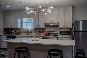 A modern kitchen with grey cabinets, a stainless steel refrigerator, a microwave, an oven, and a chandelier. There are two stools at the island. - Charming 3 Bedrooms with Spacious Living and Convenient Location to Shopping & Interstate (Lawrenceville)