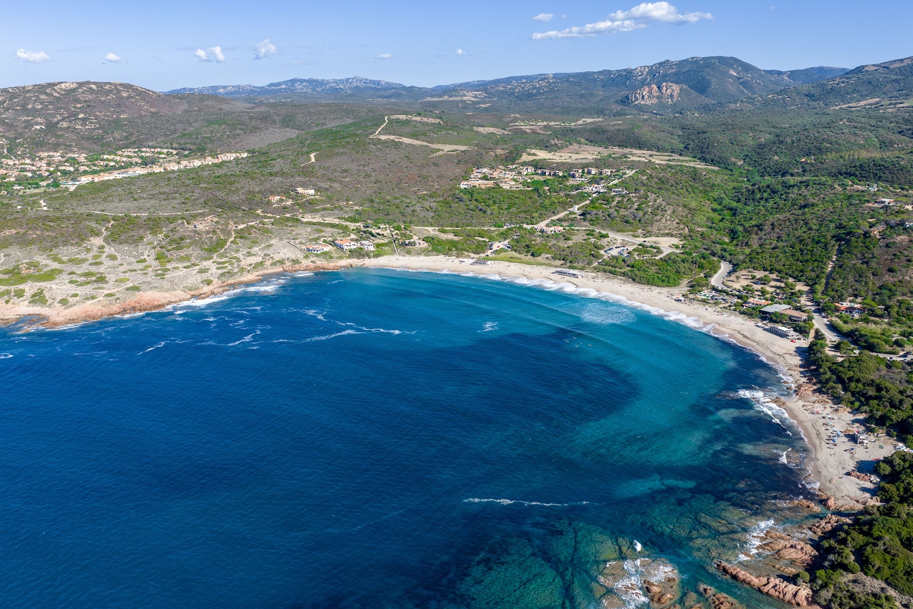 An aerial view of a coastal area with a beach, clear blue waters, and surrounding green hills.
