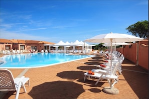 A pool area with lounge chairs and umbrellas, surrounded by buildings with tiled roofs. - Functional Studio With Shared Pool (Aglientu)