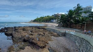 A coastal walkway with a rocky shoreline, a beach, and buildings in the background. - COOND0105 - Ondina Residence (Salvador)