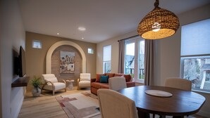 A modern living room with a dining area, featuring a large dining table, comfortable seating, a decorative wall, and a large window with curtains. - Mediterranean Suite 201 - The Agora a Boutique Hotel (Qualicum Beach)