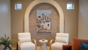 A room with two white armchairs, a small round table, a potted plant, a painting of a coastal scene, and a red cushion. - Mediterranean Suite 201 - The Agora a Boutique Hotel (Qualicum Beach)