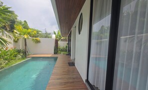 A modern outdoor pool area with a wooden deck, surrounded by lush greenery and a white wall. - Luxury 3BR Private Villa with  Pool & Bathtub Near Canggu (Dalung)