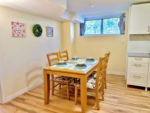 Een eethoek met een houten tafel en stoelen, een raam met uitzicht op bomen, en een keuken met witte kastjes.