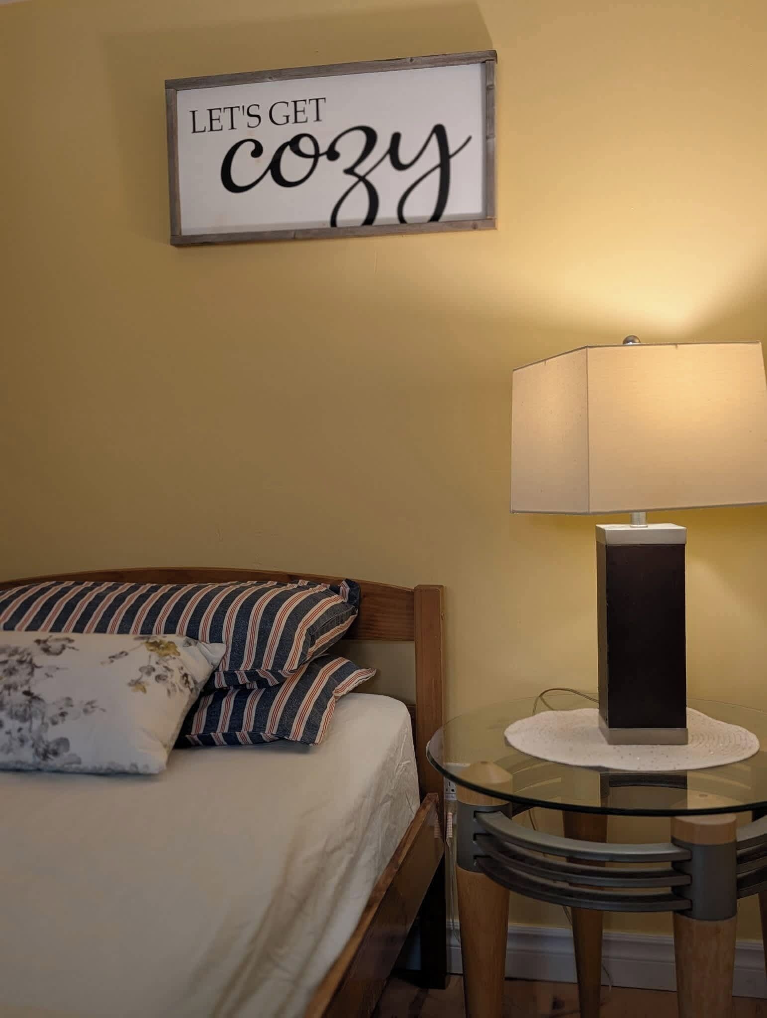 A neatly made bed with striped pillows, a bedside table with a lamp, and a sign on the wall that reads 'LET'S GET cozy'.