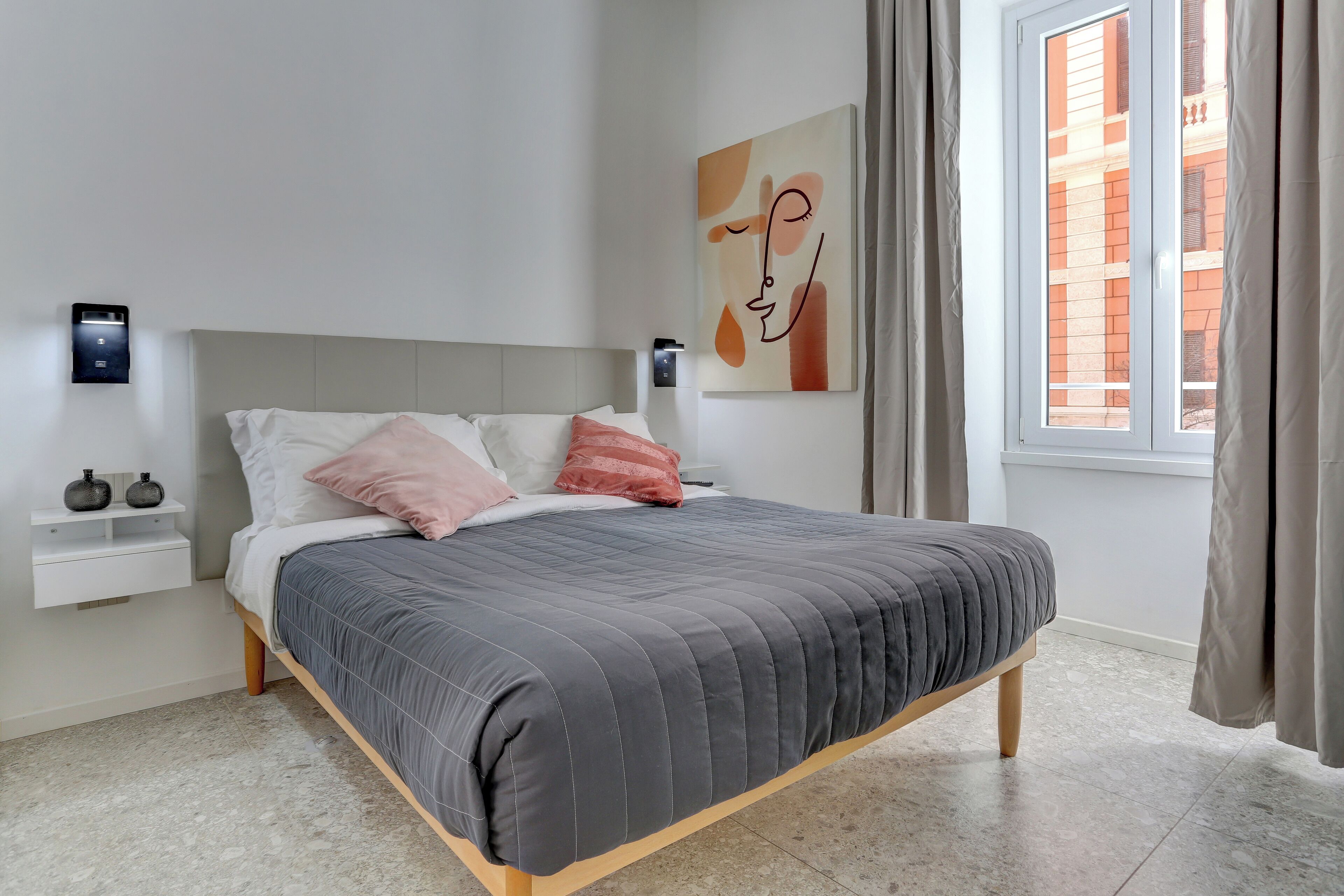 A modern bedroom with a grey quilted bedspread, a grey headboard, and a minimalist decor including a wall-mounted light and a small shelf with two vases.