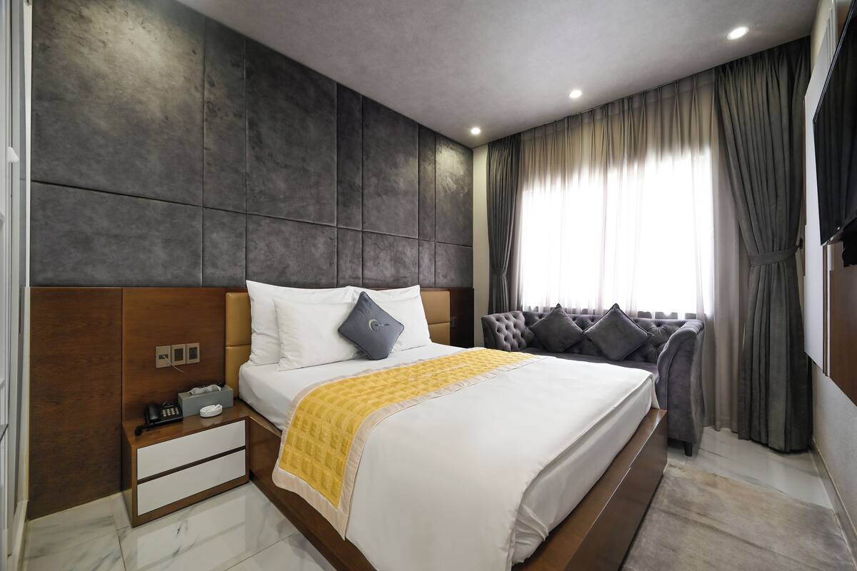 Une chambre d’hôtel moderne avec un grand lit, une table de chevet avec un téléphone et des mouchoirs, une tête de lit avec un oreiller gris et une vue sur la ville à travers de grandes fenêtres ornées de rideaux gris.
