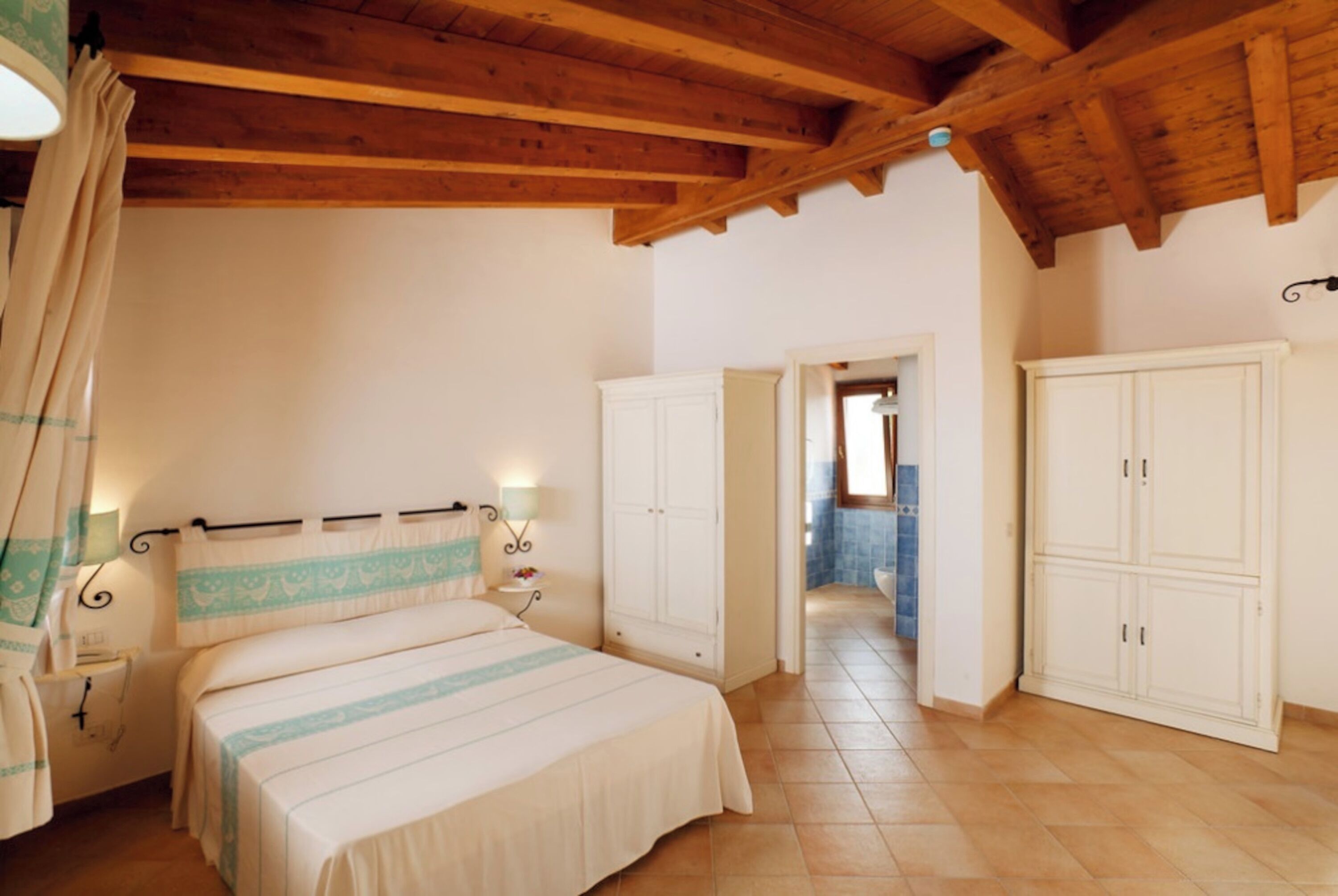Un dormitorio con cama, mesitas de noche, armario y un baño visible a través de una puerta abierta.