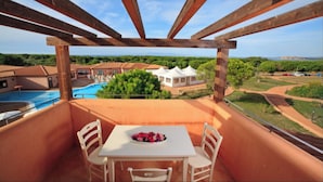 A balcony with a table and chairs, overlooking a swimming pool and a building with a red roof. - Comfy Studio With Pool (Aglientu)