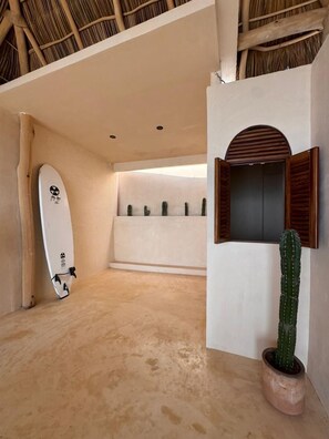 A room with a surfboard leaning against the wall, a cactus in a pot, and a wooden shelf with small plants. - Sea Breeze Penthouse at Conchas Chinas (Puerto Vallarta)