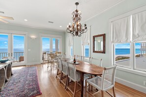 A dining area with a wooden table and white wicker chairs, large windows with ocean views, and a chandelier. - Rooftop Deck, Paddle Boards, Gulf Front on 30A (Inlet Beach)