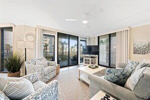 A spacious living room with a large flat-screen TV, comfortable seating, and a view of palm trees through large sliding glass doors. - Seaside Beach & Racquet 5214 (Orange Beach)