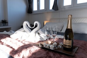 Un lit recouvert d’une couverture rose, deux serviettes en forme de cygne, une bouteille de vin et deux verres disposés sur un plateau.