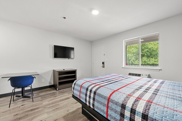 Une chambre à coucher comprenant un lit, un bureau, une chaise, un téléviseur et une fenêtre donnant sur des arbres.