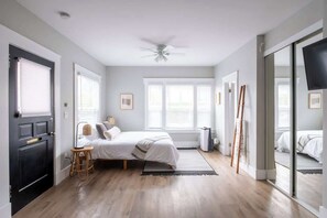 A bedroom with a bed, a chair, a standing heater, and a sliding door leading to another room. - Sunny Studio in Little Italy | Walk Everywhere (San Diego)