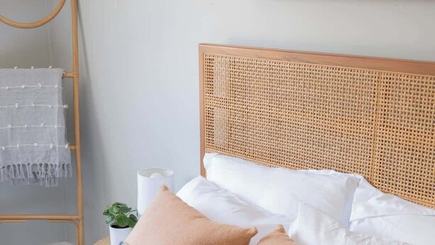 Ein Bett mit weißer Bettwäsche, zwei Kissen, einem Rattan-Kopfstück und einem Nachttisch mit Lampe und Pflanze.